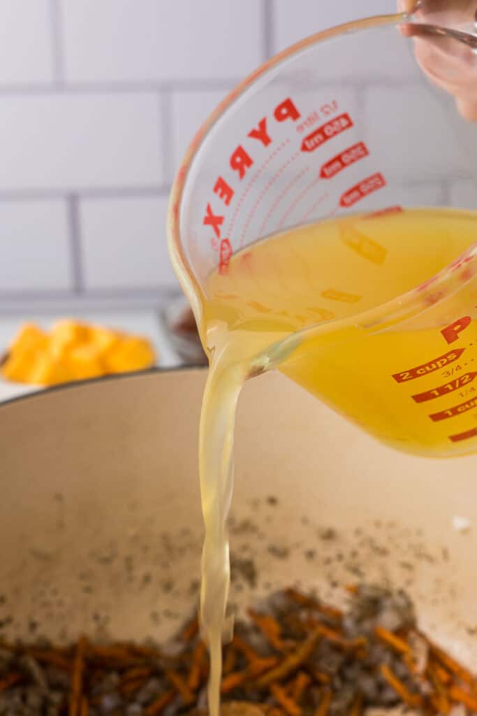 Chicken broth being added to a dutch oven with vegetables.