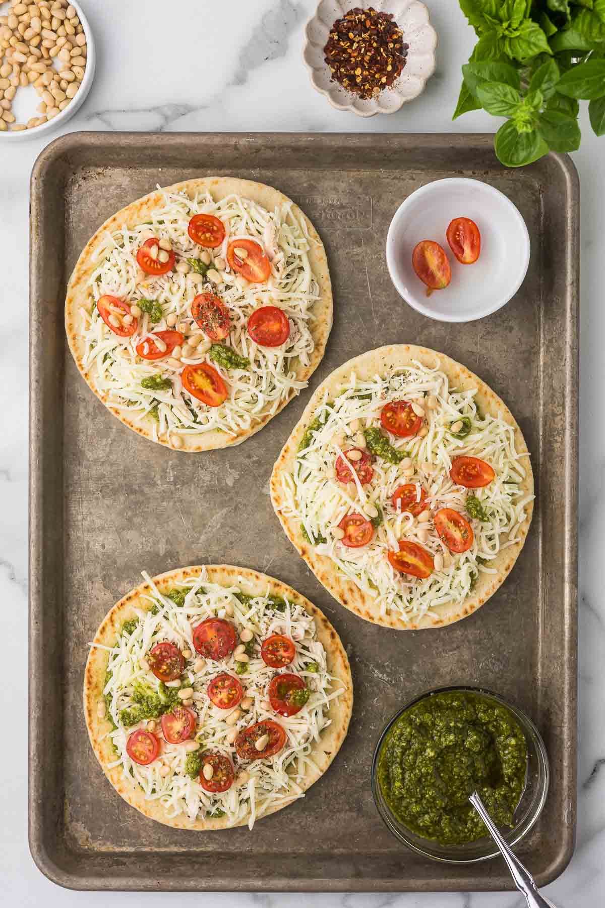 A sheet pan with three pesto chicken flatbread pizzas and a bowl of tomatoes and pesto before baking.