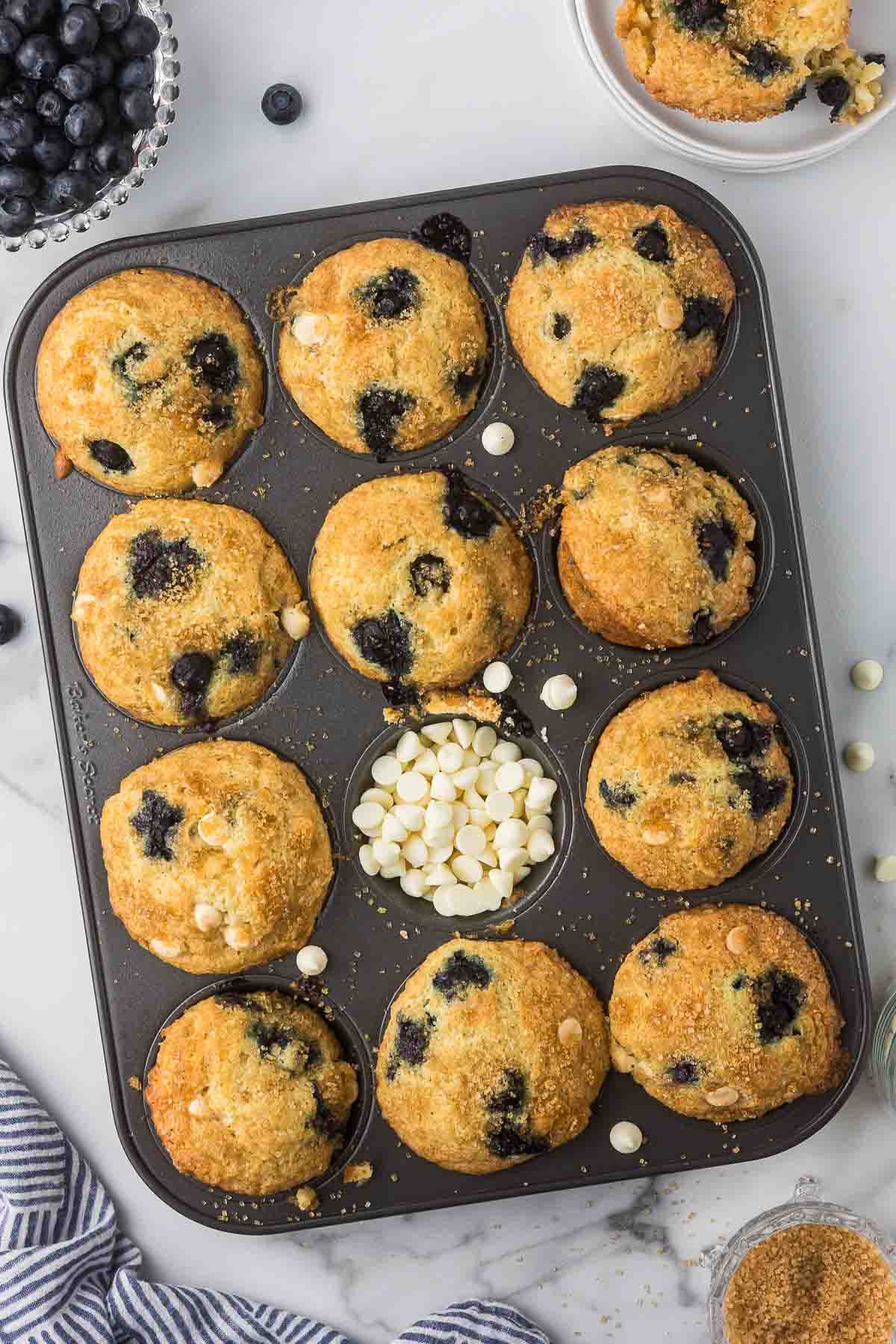 White Chocolate Blueberry Muffins in the muffin pan with blueberries in a bowl and white chocolate chips.