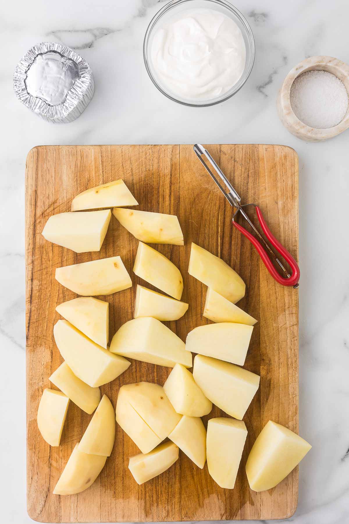 Potatoes that have been peeled and cut into quarters on a cutting board with a potato peeler.