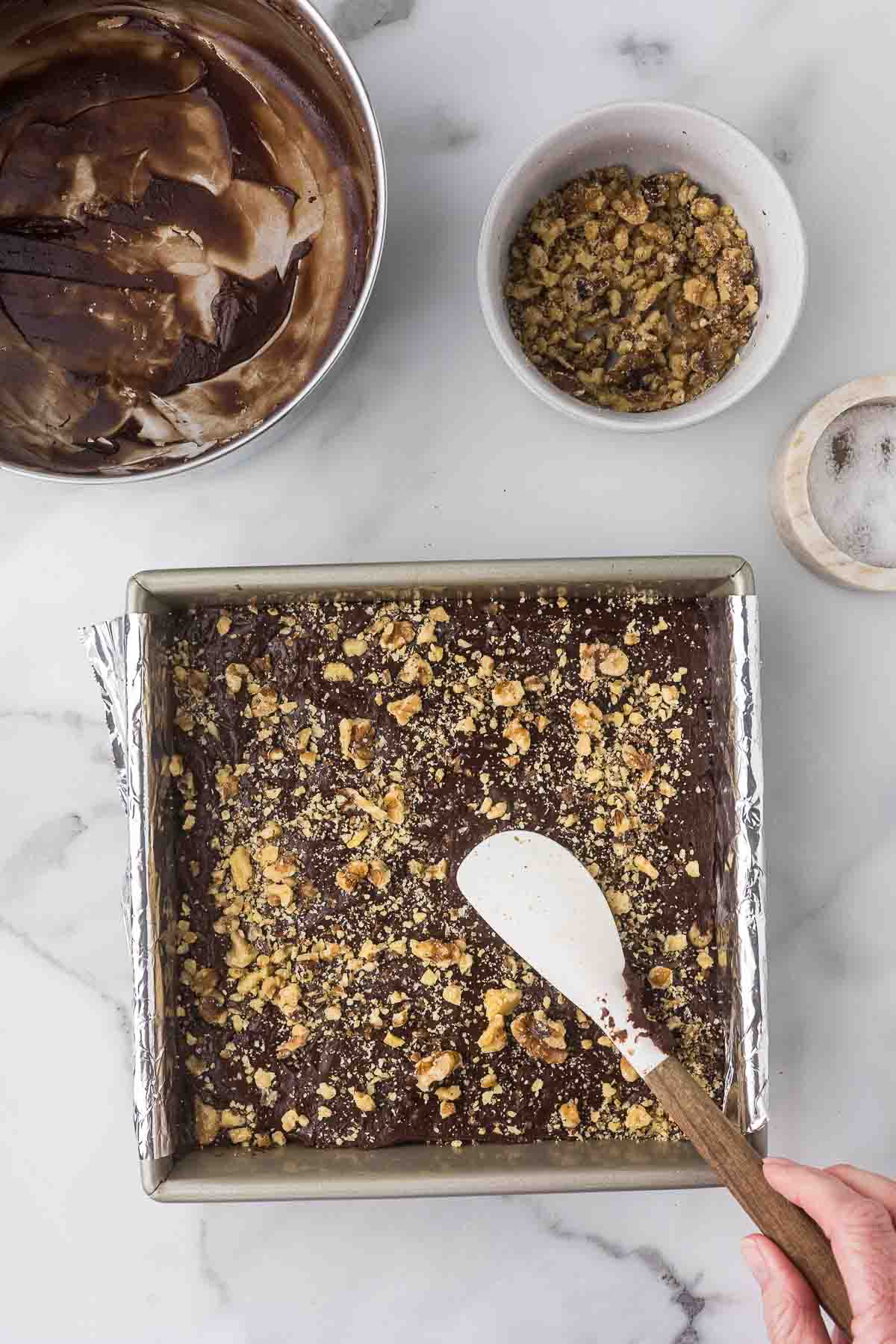 Chocolate fudge in a baking pan with a spatula patting the walnuts into the top before refrigerating.