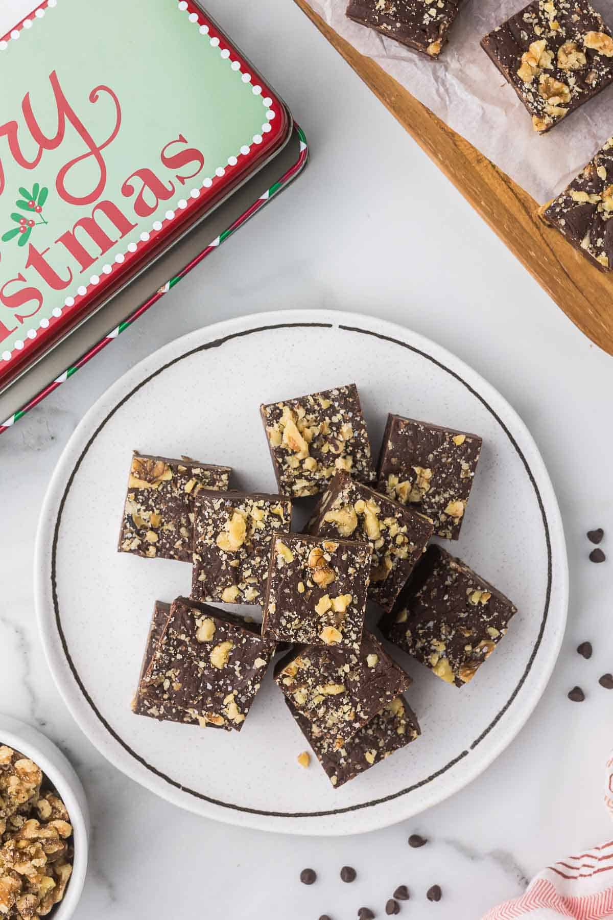 Walnut Chocolate fudge on a white plate with a Christmas tin next to the plate.