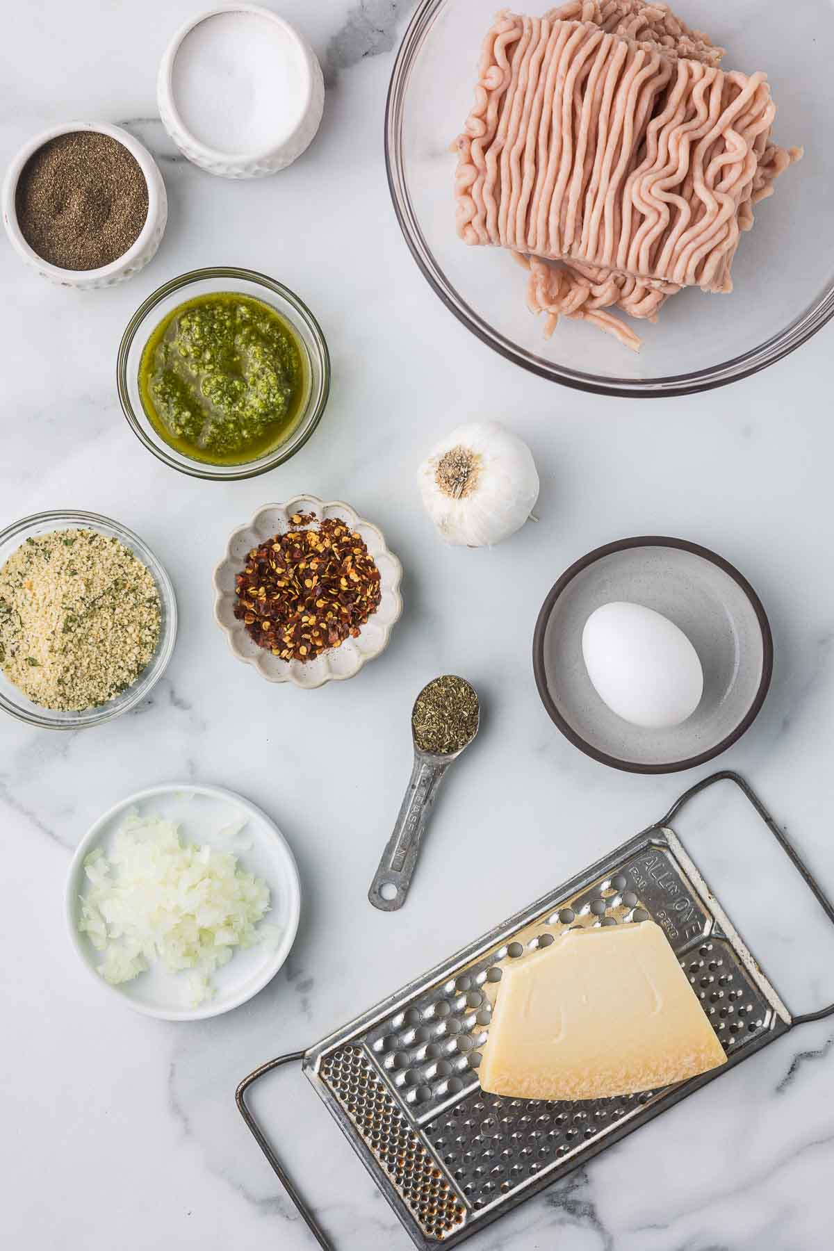 Ingredients to make pesto chicken meatballs including ground chicken, egg, parmesan cheese, pesto, red pepper flakes, garlic, bread crumbs, onion, salt and black pepper.