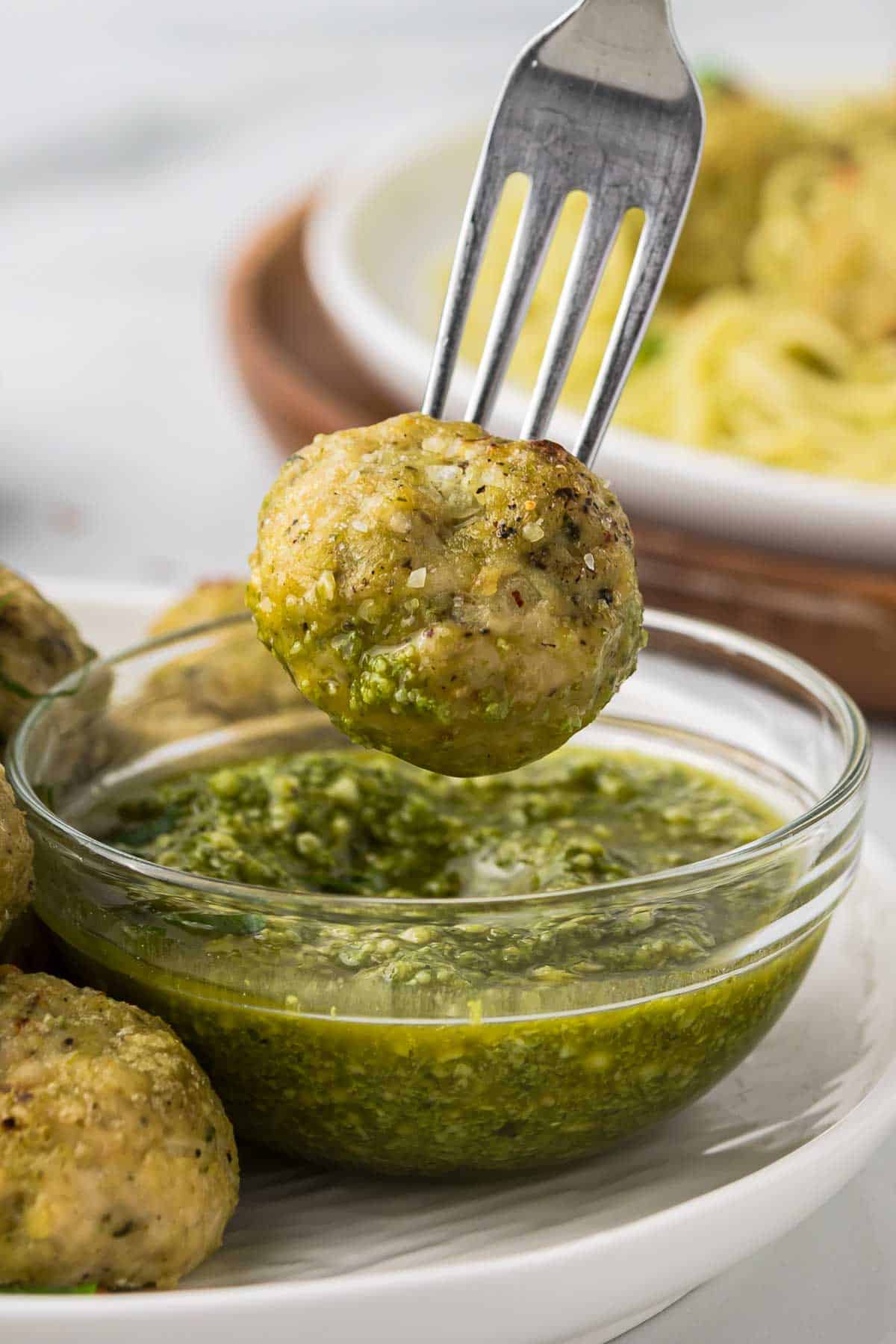 Chicken pesto meatball on a fork being dipped into pesto sauce.