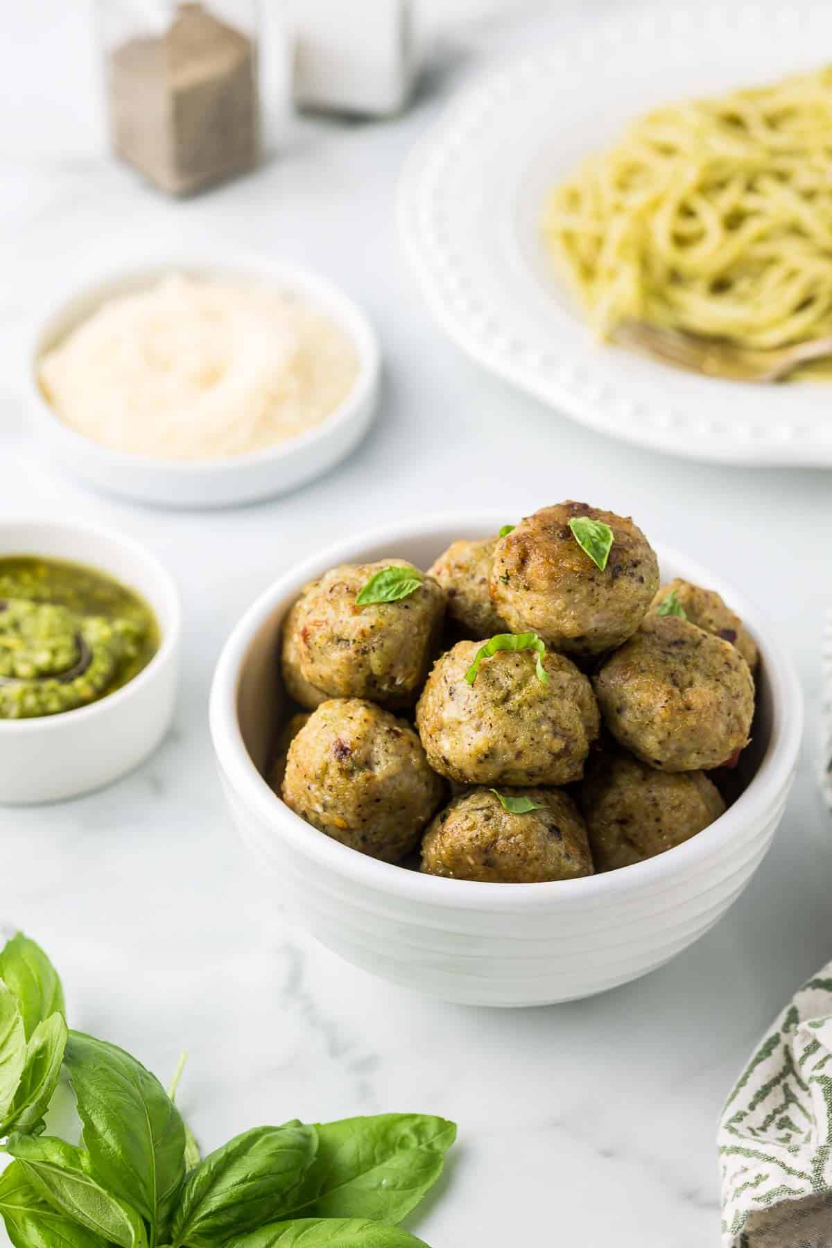 Pesto chicken meatballs in a white bowl with a plate of pasta in the back ground, a bowl of pesto, parmesan cheese and fresh basil leaves.