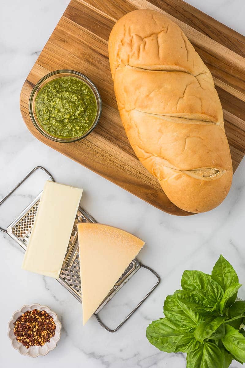 Ingredients to make pesto garlic bread including, fresh basil, red pepper flakes, bread, pesto and cheese.