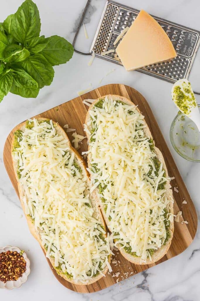 Italian bread cut length wise with pesto, parmesan cheese and mozzarella cheese on top before baking to make garlic pesto bread.