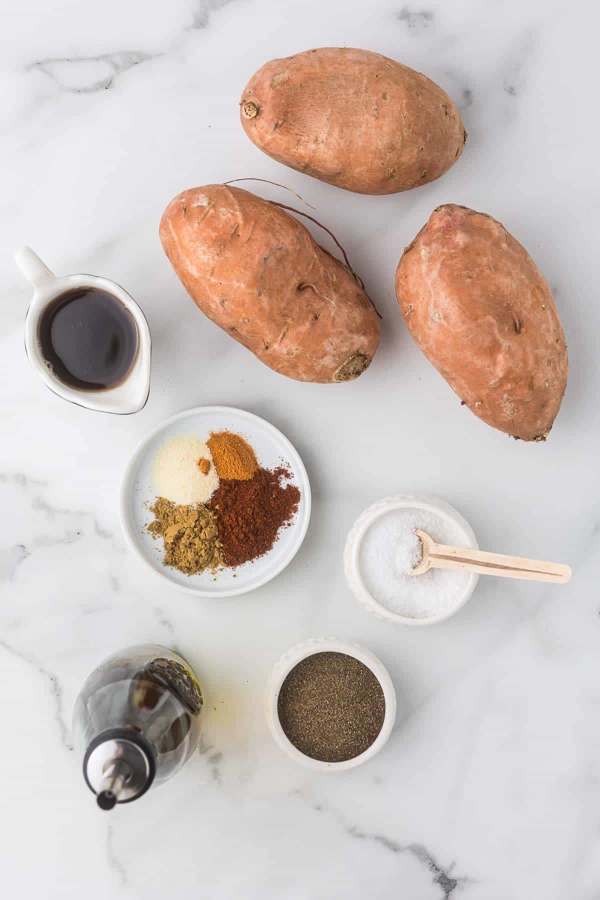 Ingredients to make roasted sweet potatoes including sweet potatoes, spices, olive oil and maple syrup to drizzle at the end.