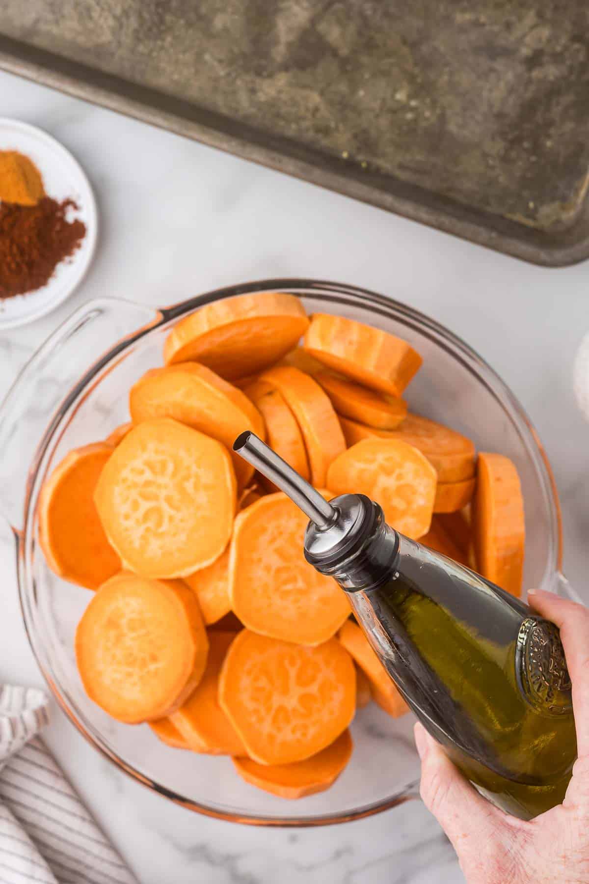 A bowl of sliced sweet potatoes in a large bowl with olive oil being drizzled over.