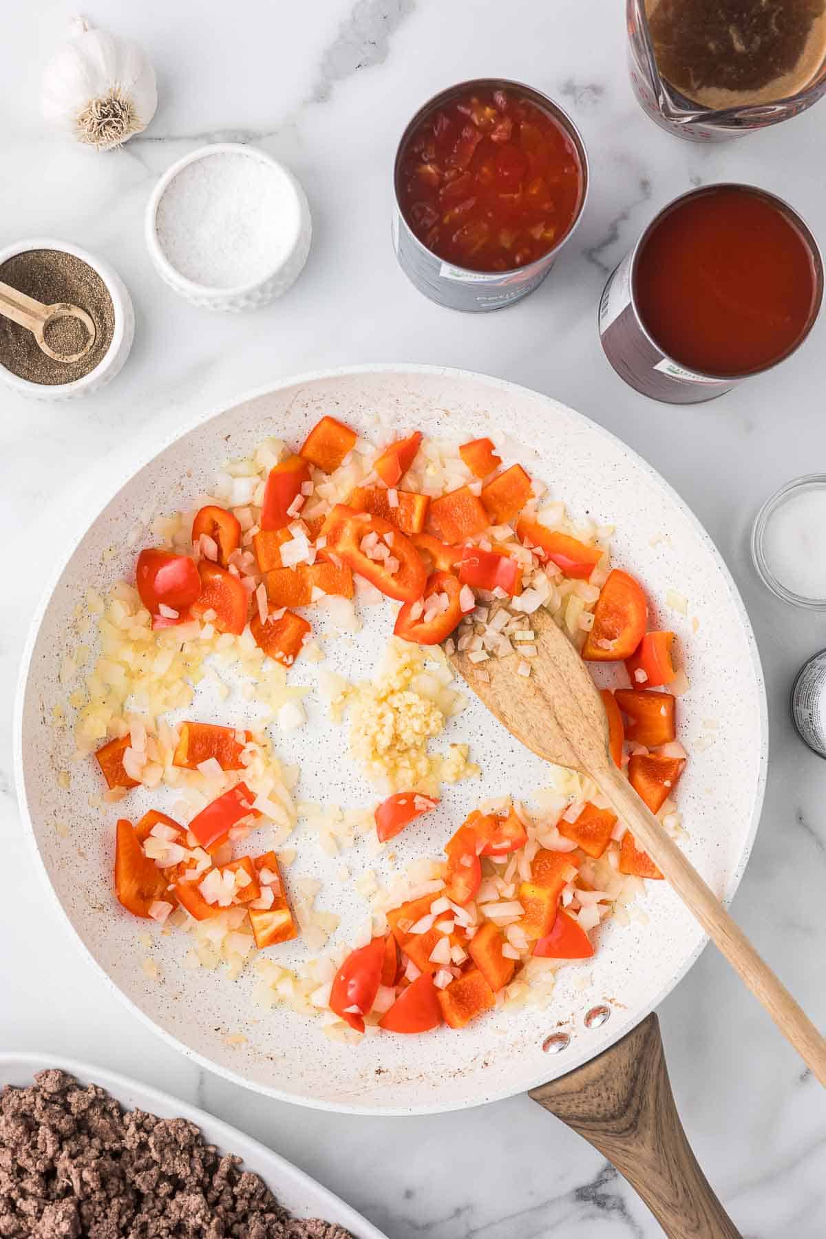 As skillet with onions, red bell peppers and garlic with a wooden spatula.