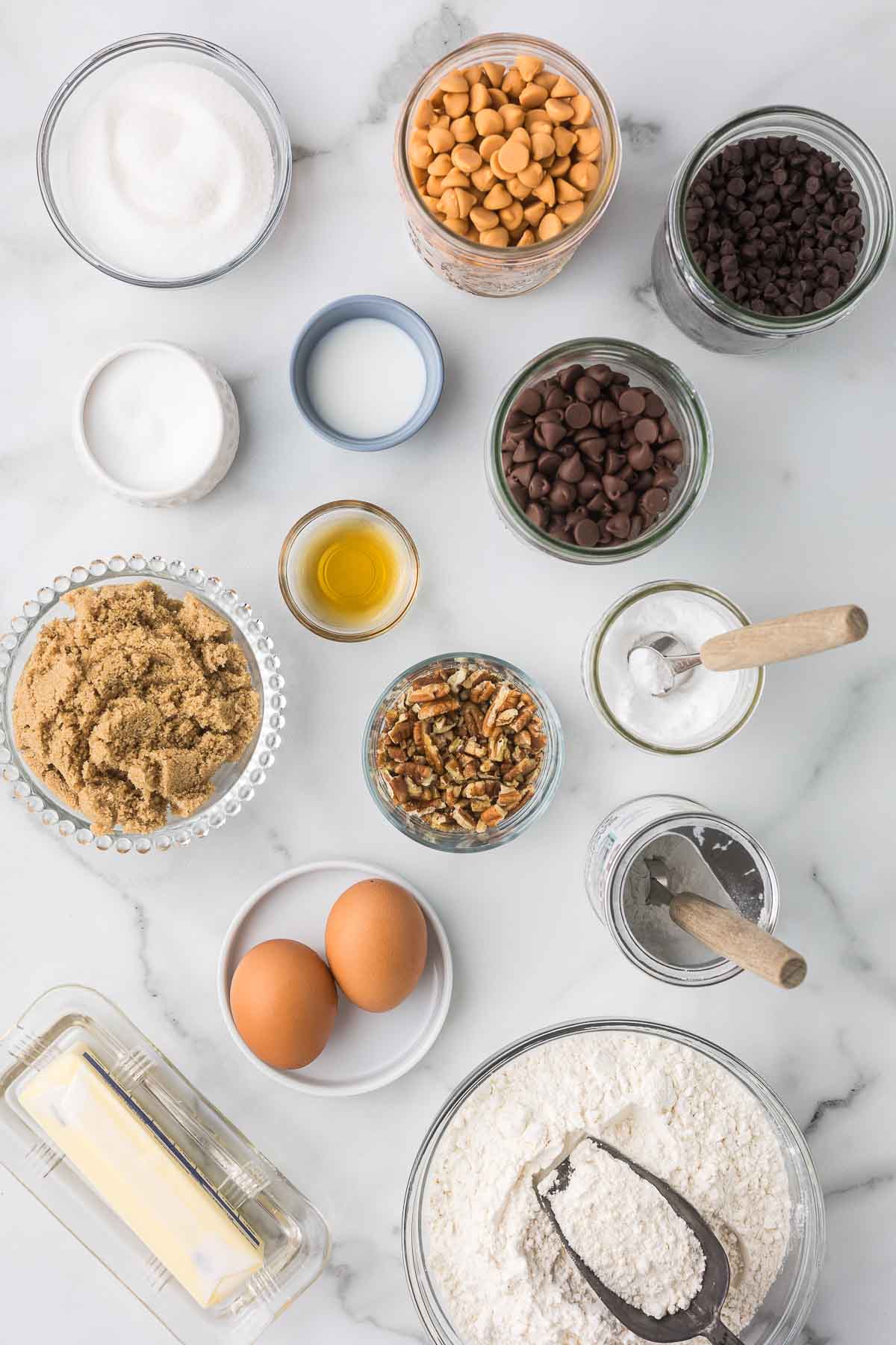 An assortment of baking ingredients on a marble surface, including brown and white sugars, butterscotch and chocolate chips, pecans, vanilla, butter, eggs, baking soda, salt, and flour.