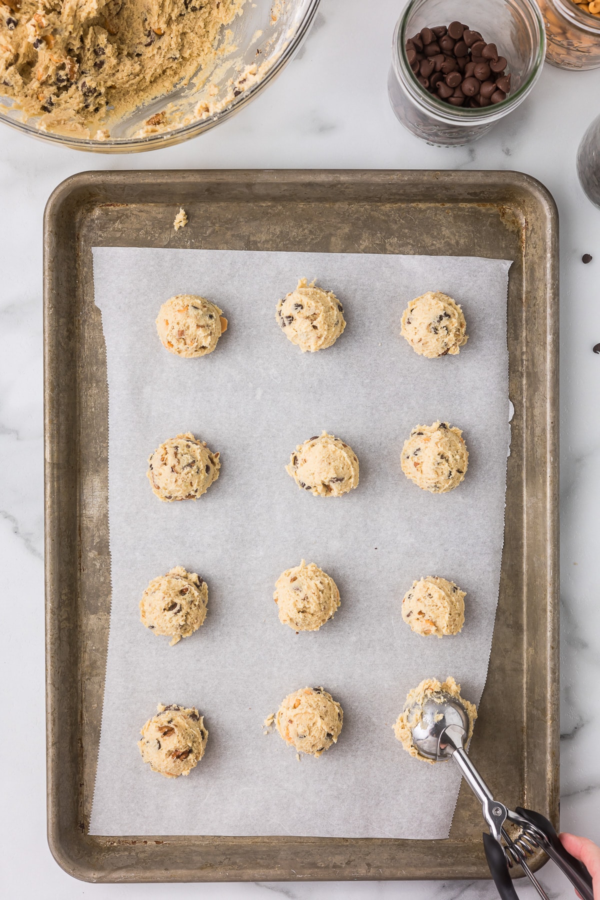 Baking tray with parchment paper holding twelve evenly spaced scoops of chocolate chip cookie dough. A hand with a scoop is placing dough on the tray. Bowls of dough and chocolate chips are in the background.