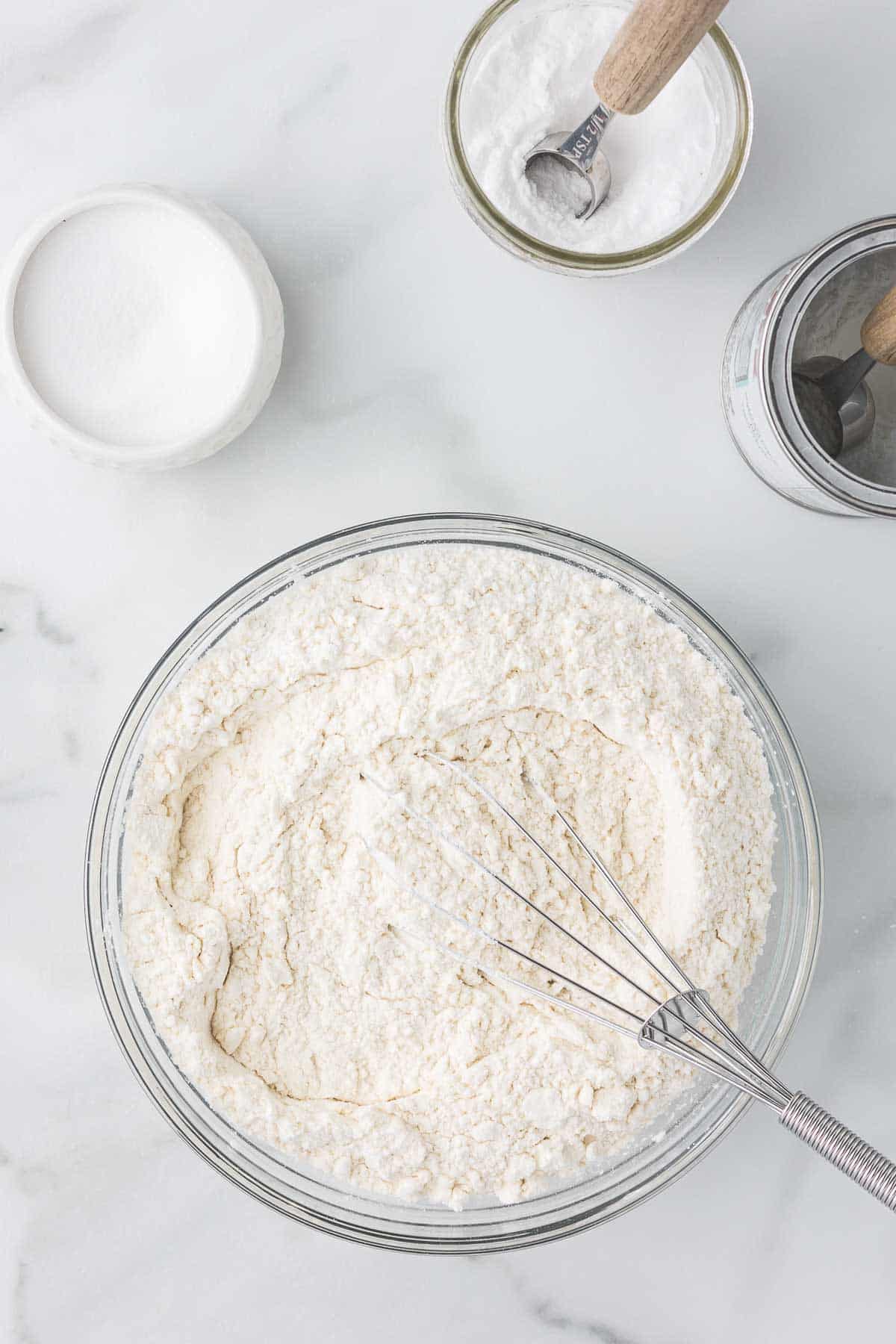 A glass bowl with flour is being whisked, surrounded by small containers of baking powder and sugar on a white marble surface. A metal measuring spoon rests in the baking powder container.