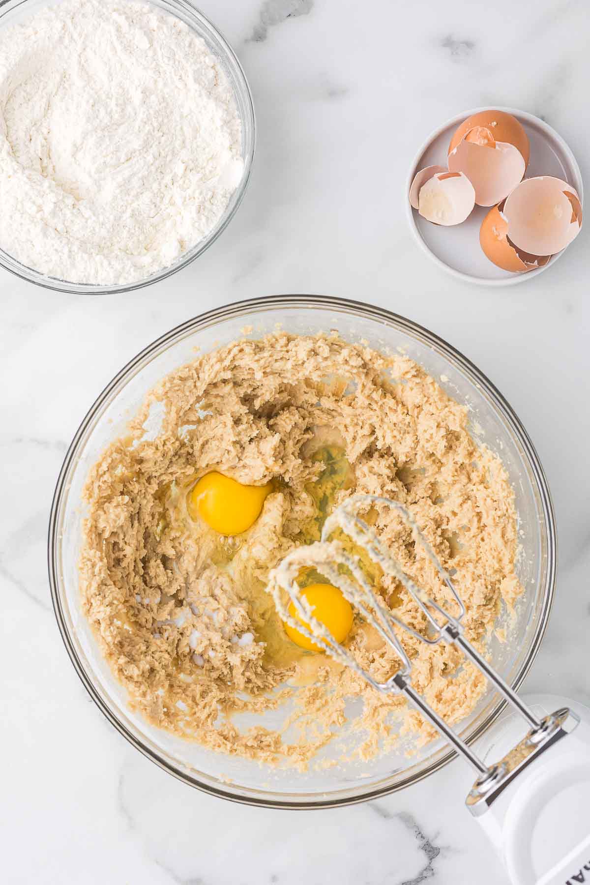 A bowl with cookie dough mixture and two eggs sits on a marble countertop. An electric mixer with beaters is partially visible. Nearby is a bowl of flour and a plate with cracked eggshells.