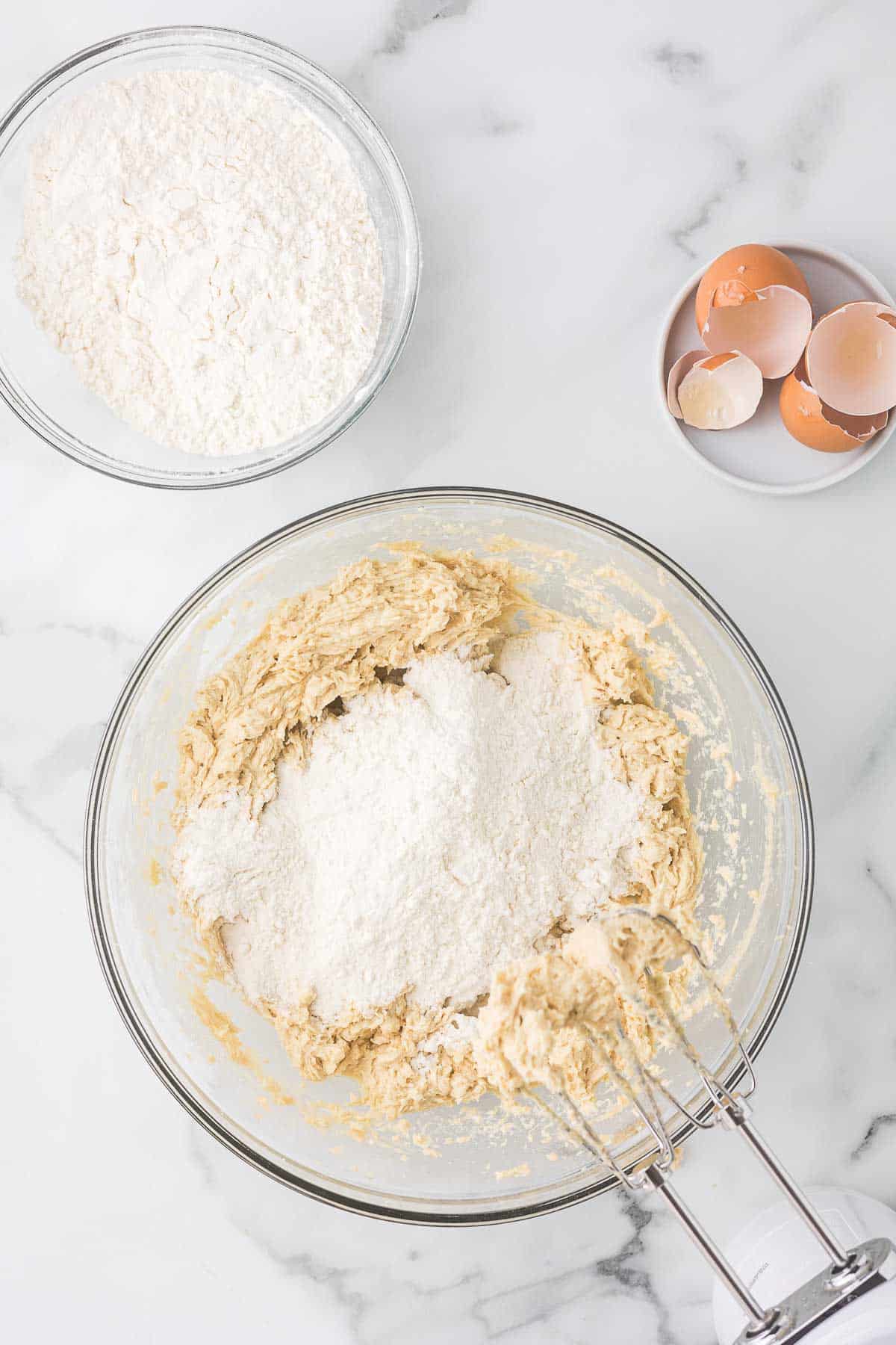 A mixing bowl with cookie dough and flour being blended with a hand mixer, next to a bowl of flour and a small plate with broken eggshells. All items are placed on a marble countertop.