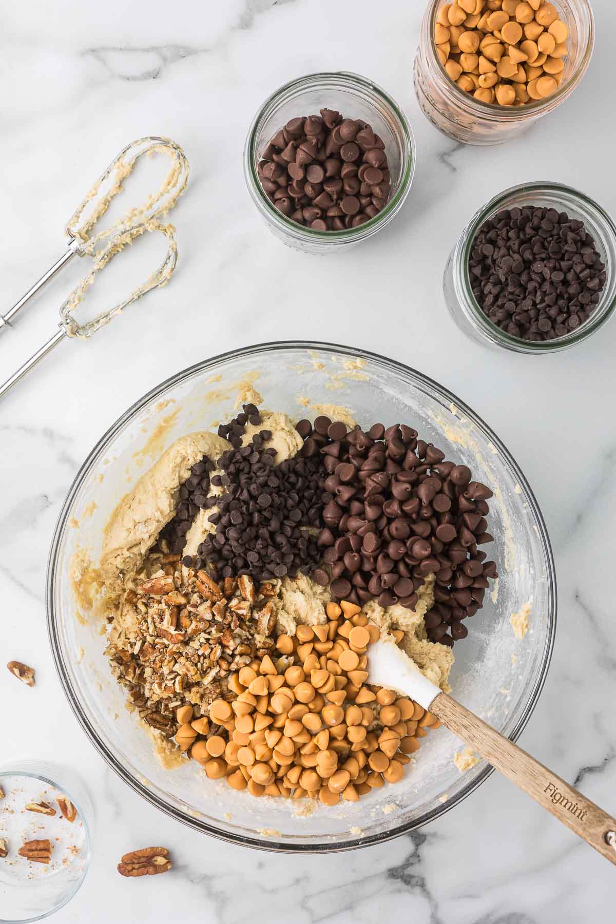 A mixing bowl filled with cookie dough, chocolate chips, butterscotch chips, and chopped nuts. Three jars with various chips, a wooden spoon, and a hand mixer are nearby on a marble countertop.