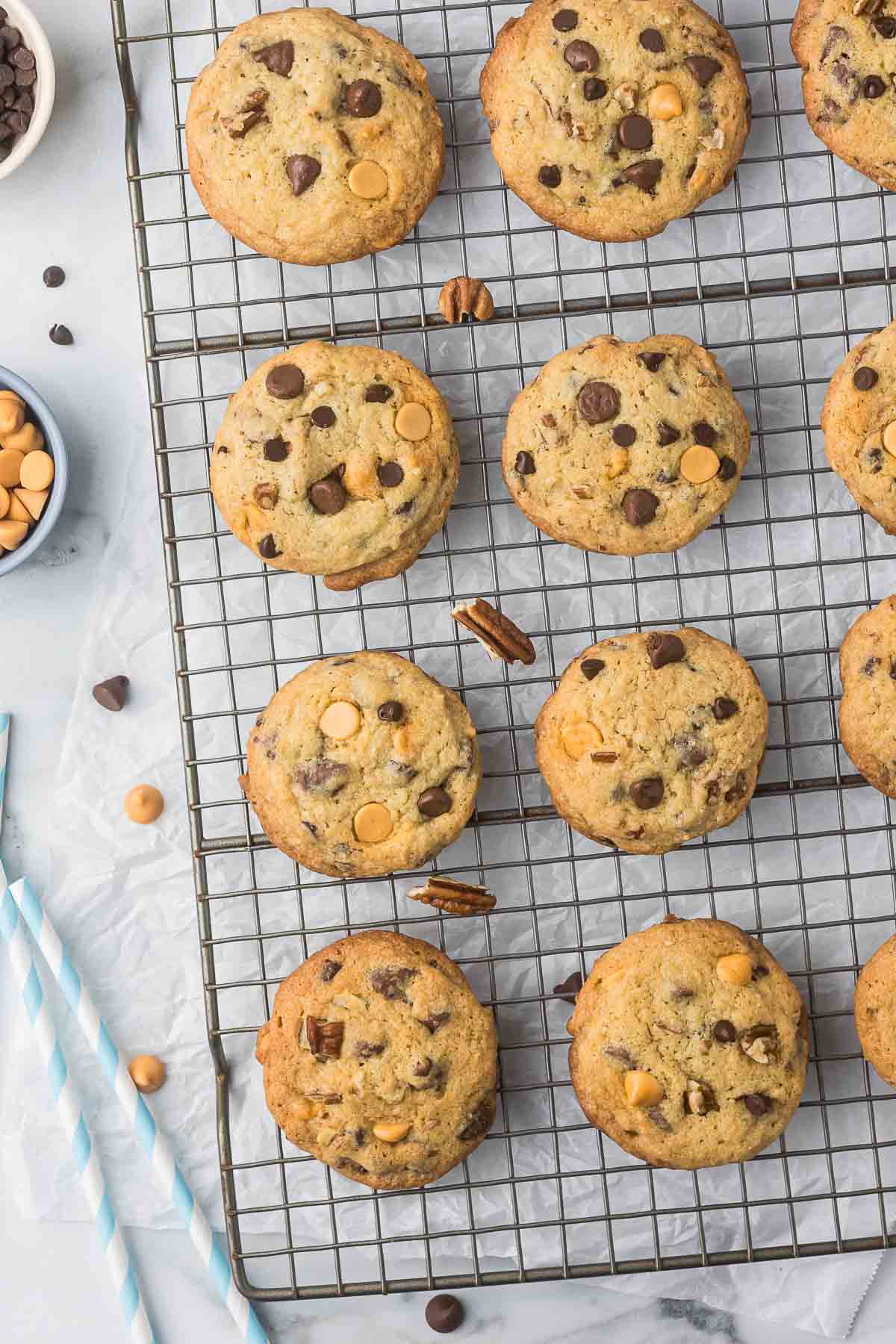 A batch of freshly baked cookies with chocolate and butterscotch chips cooling on a wire rack. The cookies are arranged neatly, and there are straws and extra chips nearby on the parchment paper.