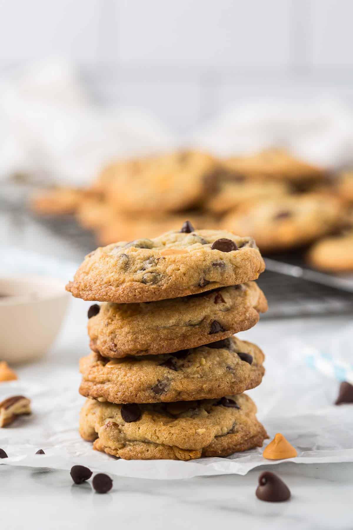 A stack of four chocolate chip cookies on a piece of parchment paper. More cookies are visible in the background, slightly out of focus, on a cooling rack. Scattered chocolate and butterscotch chips are around the cookies.