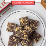 A plate of square-shaped chocolate walnut fudge pieces stacked neatly, surrounded by a cookbook and festive decorations. The image includes a text overlay with the delicious recipe source: cookingupmemories.com.