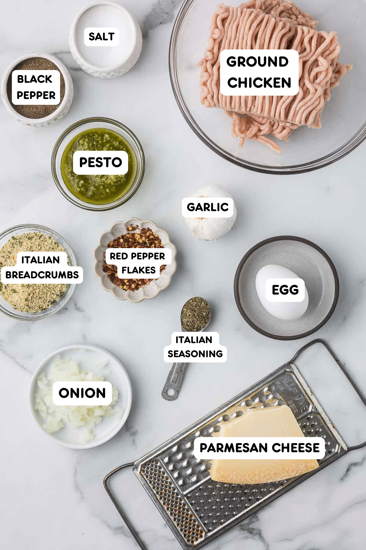 On a marble countertop, ingredients are labeled: ground chicken, pesto, salt, black pepper, garlic, Italian breadcrumbs, red pepper flakes, Italian seasoning, onion, egg, and Parmesan cheese with a grater. Nearby sits a plate of chocolate chip butterscotch cookies for dessert inspiration.