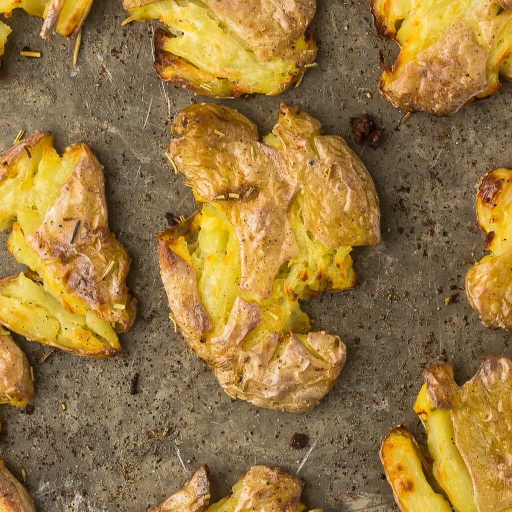 Close-up of crispy smashed potatoes on a baking sheet. The golden potatoes have a rustic appearance and are seasoned with visible bits of herbs. The uneven texture and browning suggest they are freshly roasted.