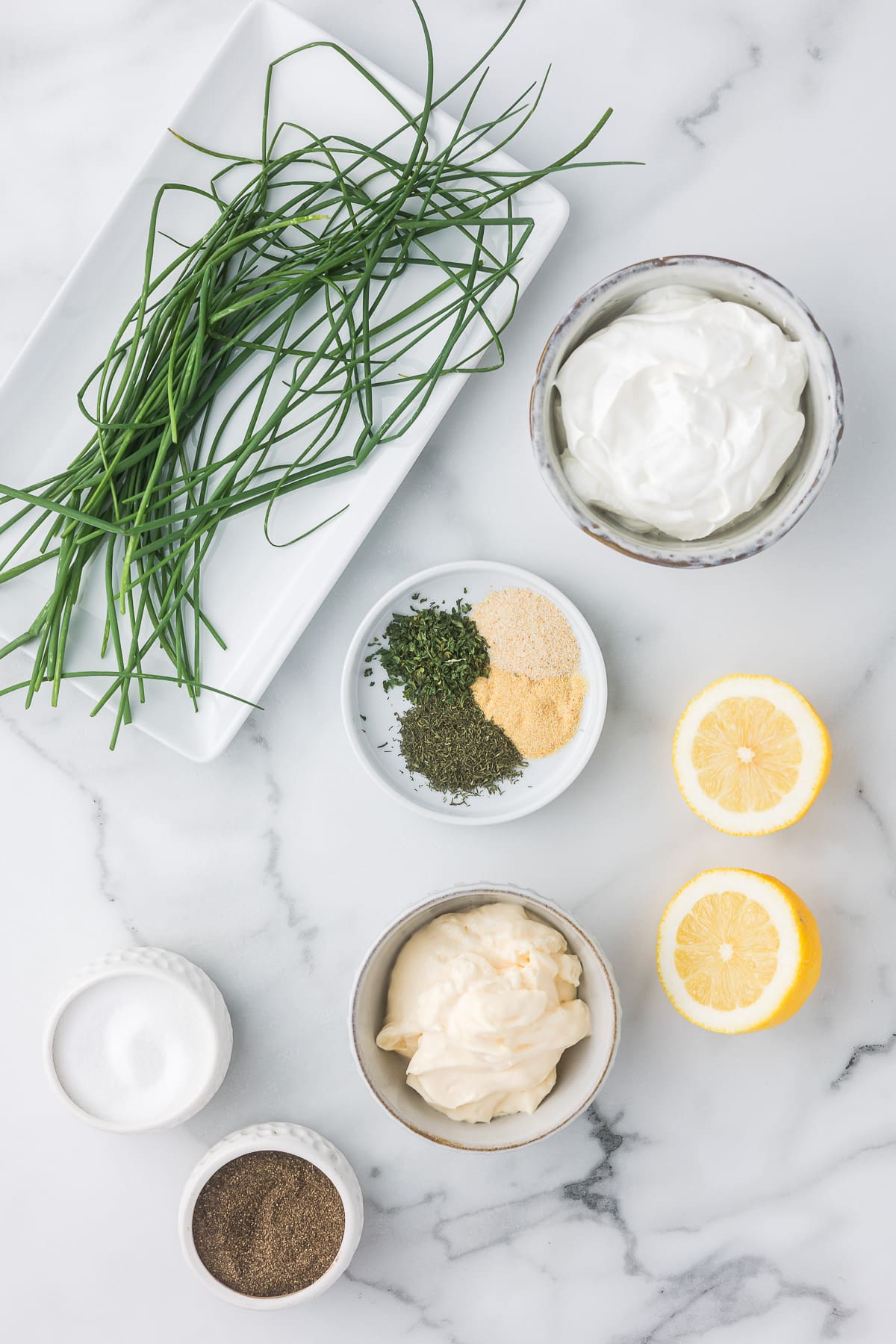 Ingredients on a marble surface: fresh chives on a rectangular plate, bowls of sour cream, mayonnaise, dried herbs, garlic powder, lemon halves, and small bowls of salt and pepper.