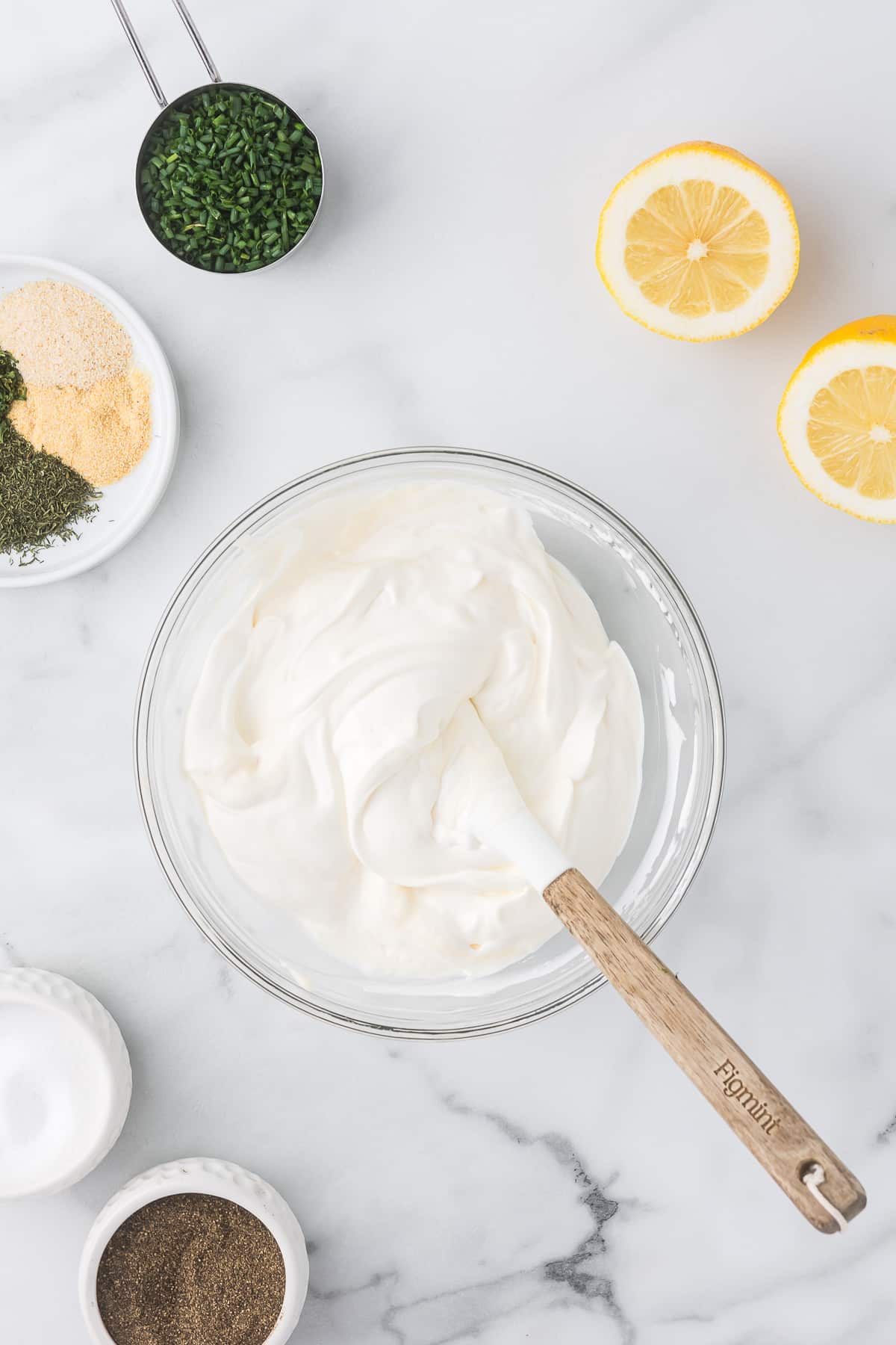 A bowl of creamy white dip with a wooden spatula on a marble surface. Nearby are a measuring cup of chopped herbs, halved lemons, and small bowls of powdered spices and pepper.