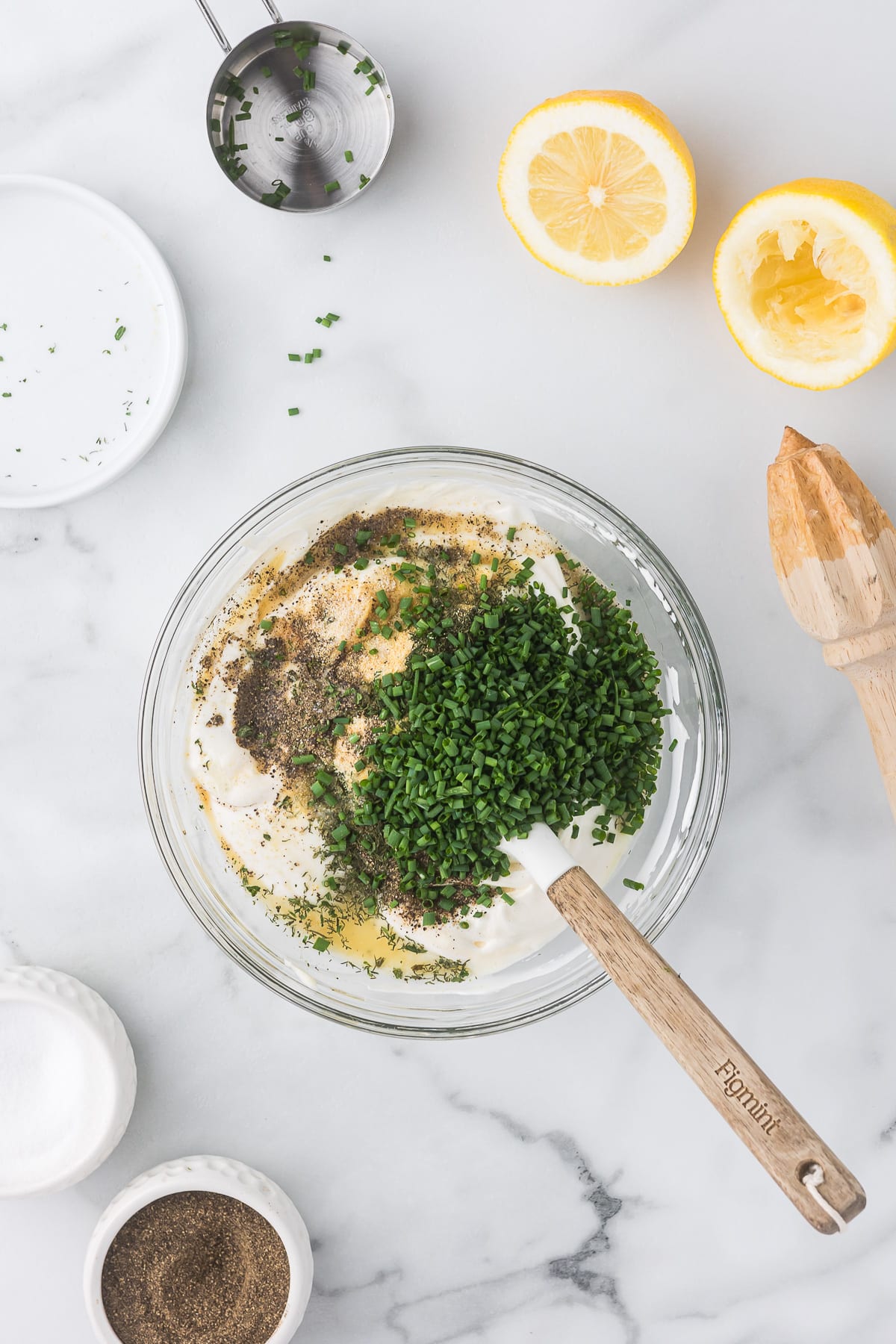 A glass bowl filled with a creamy mixture topped with chopped chives and spices sits on a marble surface. Nearby are halved lemons, a wooden citrus juicer, a small bowl of pepper, and a measuring cup.