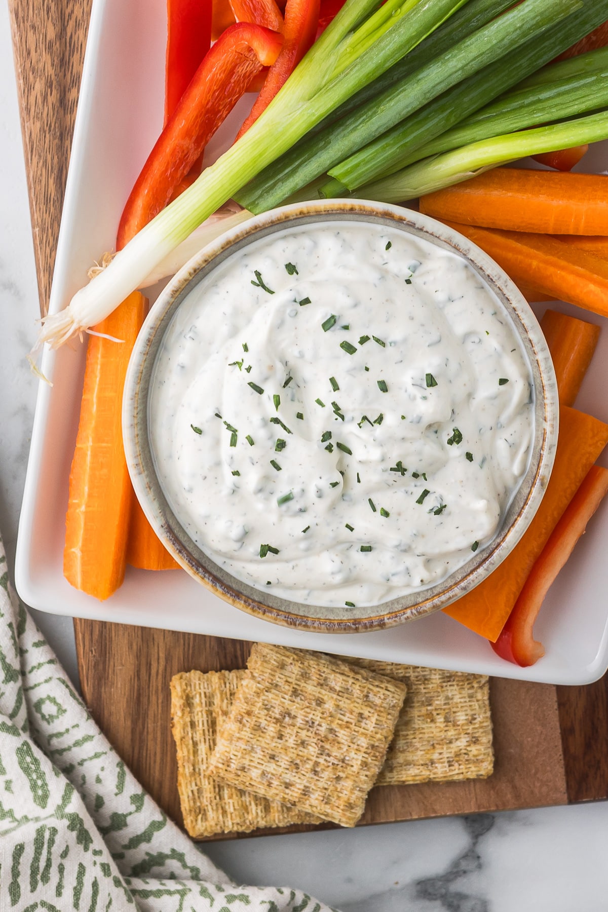 A bowl of creamy white dip garnished with herbs is surrounded by fresh vegetables, including carrot sticks, red bell pepper slices, and green onions, on a white platter. Three square crackers are placed beside the vegetables.