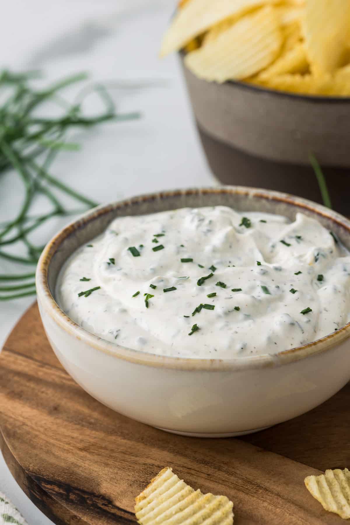 A bowl of creamy chive dip garnished with chopped herbs sits on a wooden board. In the background, a bowl of ridged potato chips is visible, placed on a light surface with scattered green chives.