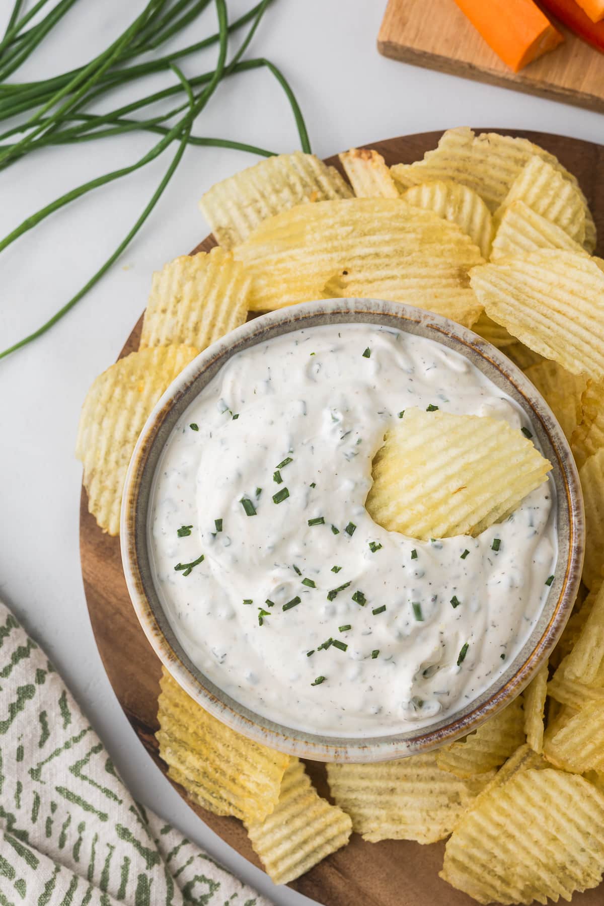 A bowl of creamy sour cream and chive dip garnished with chopped herbs sits surrounded by crinkle-cut potato chips on a wooden platter. Fresh chives are visible in the background, and a patterned cloth and vegetable sticks are partially visible nearby.