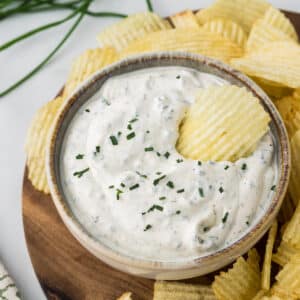 A bowl of creamy ranch dip topped with chopped chives sits on a wooden board surrounded by wavy potato chips, with one chip dipped into the ranch.