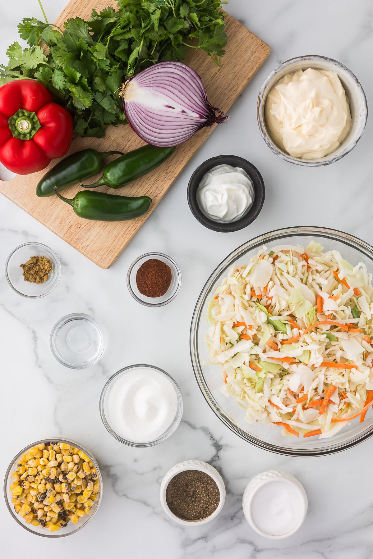 Top view of coleslaw ingredients: chopped cabbage and carrots in a bowl, red pepper, cilantro, red onion, and jalapeños on a board. Small bowls contain corn, mayonnaise, sour cream, spices, and sugar, all on a marble countertop.