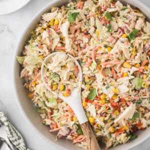 A large bowl filled with creamy chicken salad, featuring shredded chicken, corn, diced bell peppers, carrots, and cilantro. A wooden spoon rests in the salad. The background includes a patterned cloth napkin.