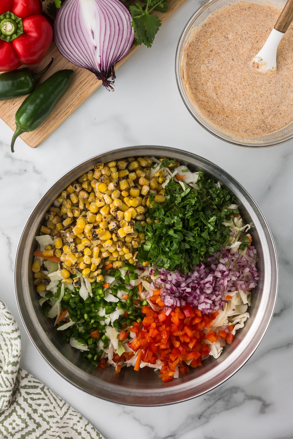 A bowl filled with chopped corn, cilantro, red onion, jalapeño, and red bell pepper sits on a marble countertop next to a bowl of seasoning mix. A cutting board with more vegetables is nearby.