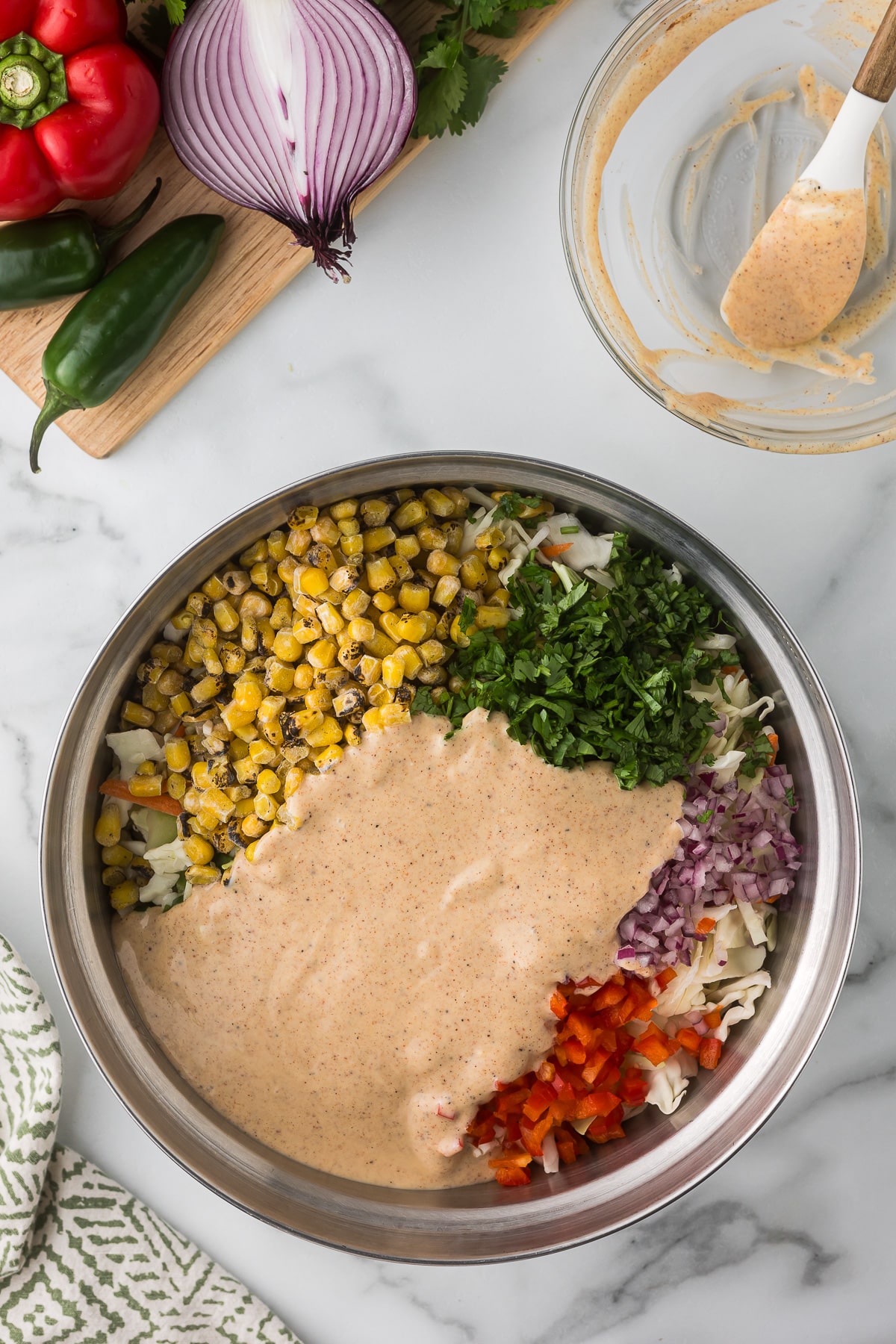 A large metal bowl filled with diced red onions, roasted corn, chopped cilantro, and red bell peppers, covered with a creamy sauce. Beside it, a cutting board with a halved red onion, a jalapeño, a red bell pepper, and an empty glass bowl.