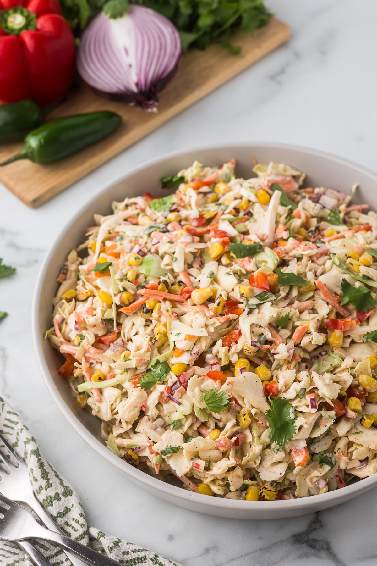 A bowl of creamy coleslaw filled with chopped cabbage, red bell peppers, corn, and cilantro on a marble countertop. In the background, a cutting board with red onion, red bell pepper, jalapeño, and cilantro.