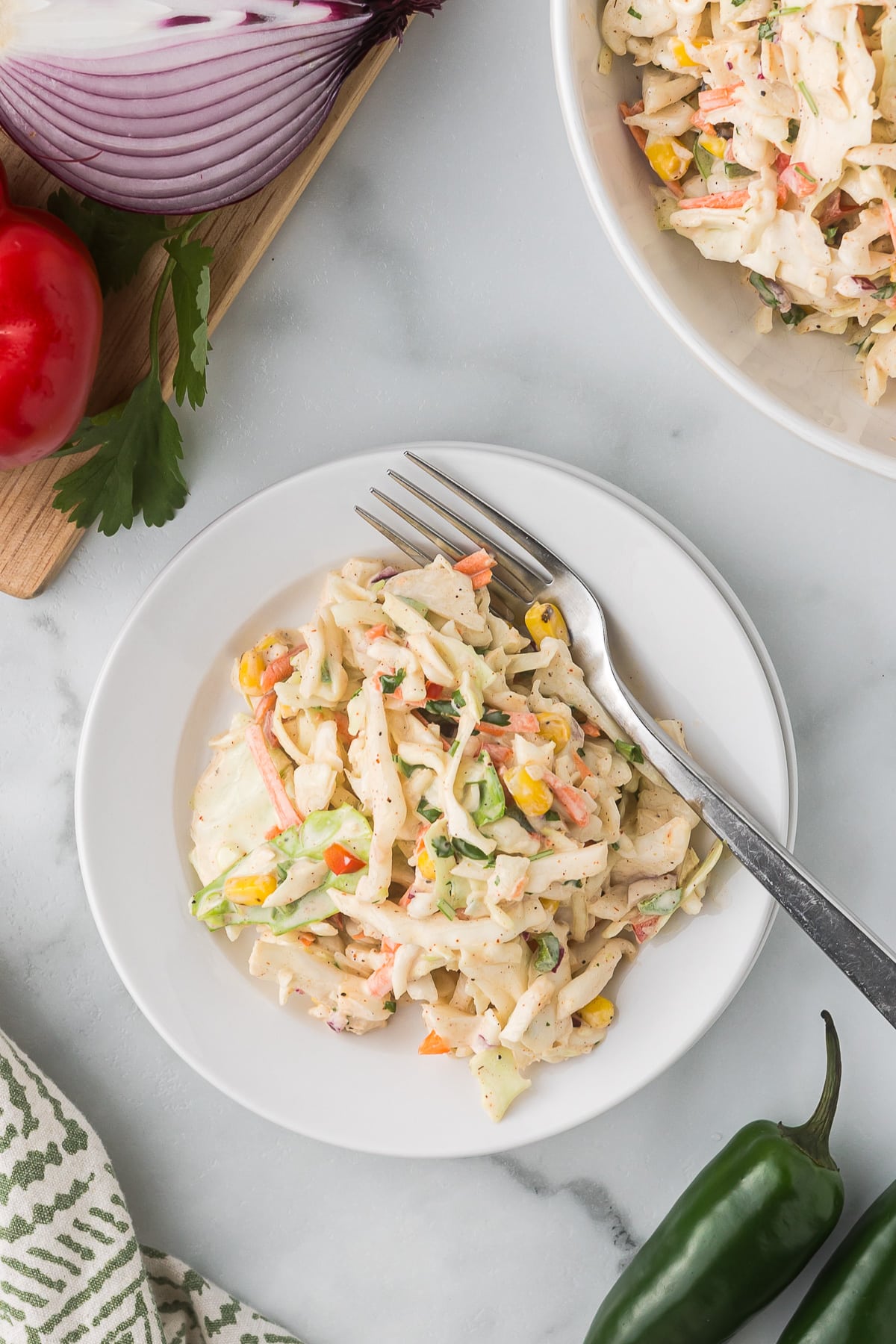 A plate of creamy coleslaw with shredded cabbage, carrots, and corn, garnished with herbs. A fork rests on the plate. Nearby are a half red onion, a tomato, a jalapeño, and a cutting board with parsley on a marble surface.