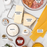 Top-down view of lasagna ingredients on a marble surface: ground beef, Italian sausage, mozzarella, Parmesan, ricotta, salt, pepper, red pepper flakes, oregano, sweet basil, garlic, tomato paste, onion, chicken broth, and lasagna noodles.