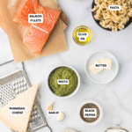 Top-down view of various ingredients on a marble surface, including labeled salmon fillets, pasta, red pepper flakes, olive oil, butter, pesto, Parmesan cheese, garlic, black pepper, salt, and heavy cream. A cheese grater is also visible.