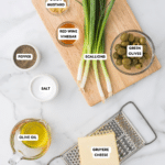 Ingredients for a recipe arranged on a marble surface: scallions on a wooden board, a bowl of green olives, grated Gruyere cheese on a grater, olive oil in a pitcher, and small bowls of Dijon mustard, red wine vinegar, pepper, and salt.