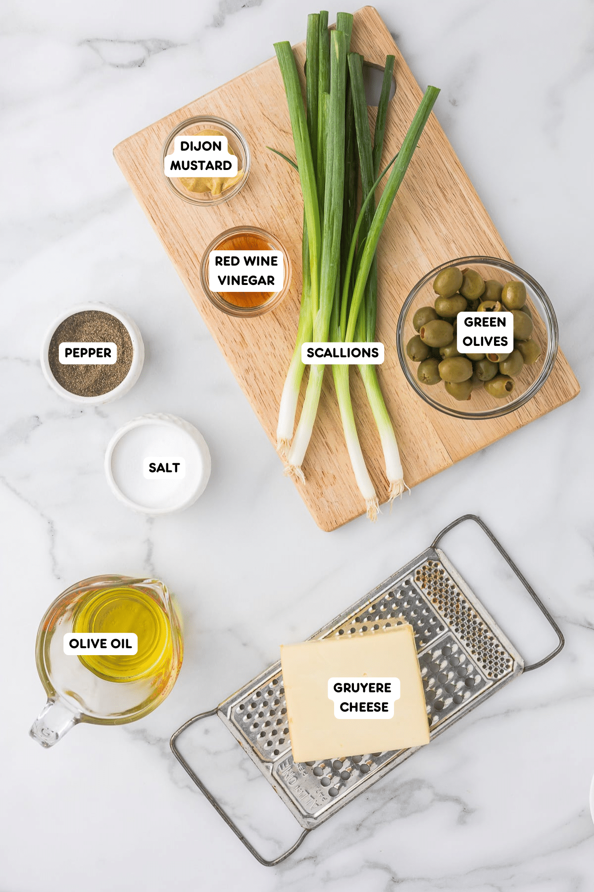 Ingredients for a recipe arranged on a marble surface: scallions on a wooden board, a bowl of green olives, grated Gruyere cheese on a grater, olive oil in a pitcher, and small bowls of Dijon mustard, red wine vinegar, pepper, and salt.