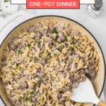 A skillet filled with creamy hamburger stroganoff topped with chopped parsley. A white serving spoon rests in the skillet. The text reads, "The Best Hamburger Stroganoff, One-Pot Dinner," with a website for the full recipe.