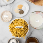 Ingredients for a dish are laid out on a marble surface: ground beef, butter, spices, egg noodles, flour, sour cream, milk, and beef broth. Each ingredient is placed in separate bowls or containers, labeled with text.