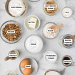 Top-down view of baking ingredients on a marble surface, including sour cream, pecans, sugar, butter, powdered sugar, flour, eggs, vanilla, ground cinnamon, milk, baking soda, and baking powder, each labeled in its own container.