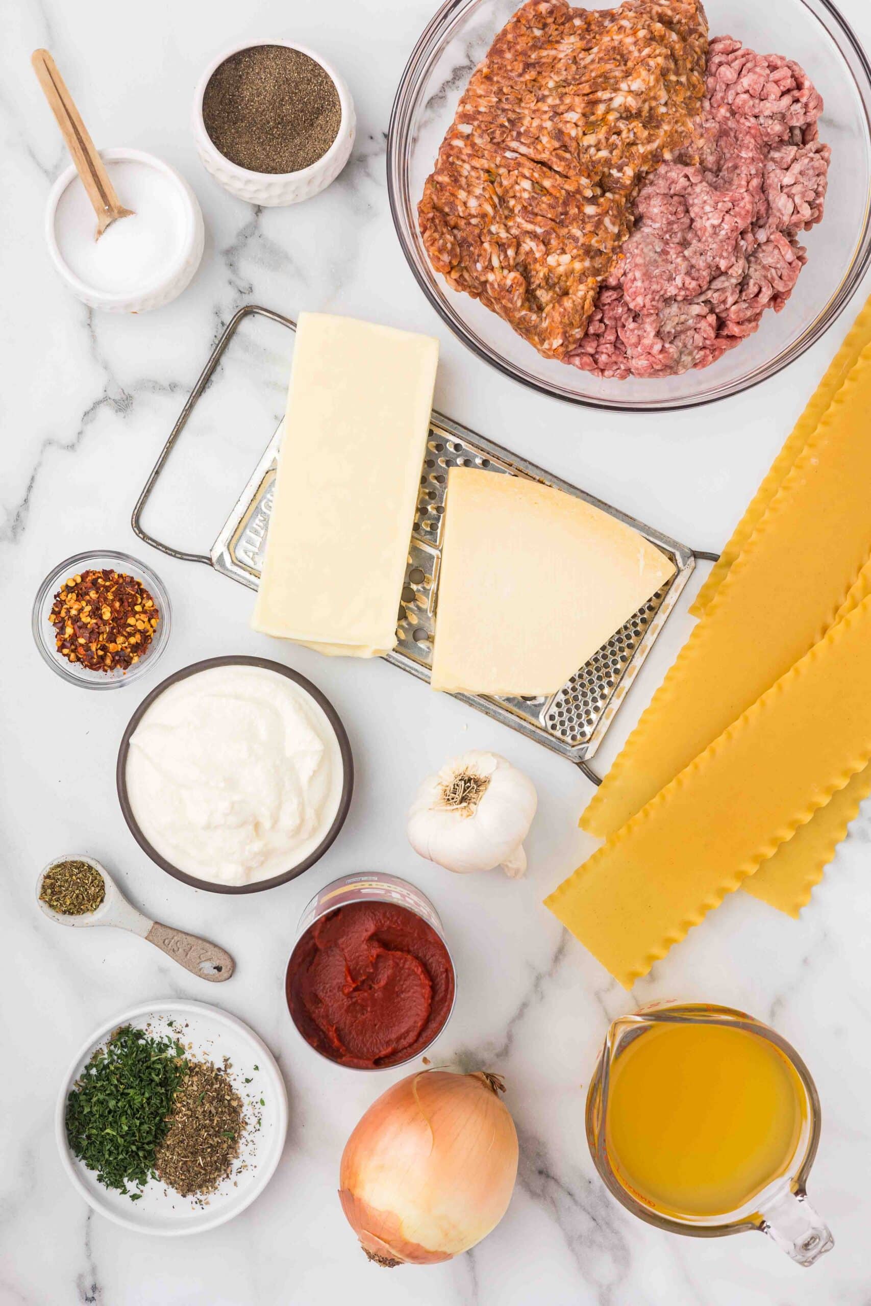 Ground beef and sausage in a glass bowl, a bowl of pepper, a bowl of salt with a spoon, cheese grater with a block of parmesan and mozzarella on it, lasagna noodles, bowl of red chili flakes, bowl of ricotta cheese, bulb of garlic, bowl of tomato paste, an onion, a measuring cup full of broth, a teaspoon of pepper, and a shallow dish of basil and black pepper. 