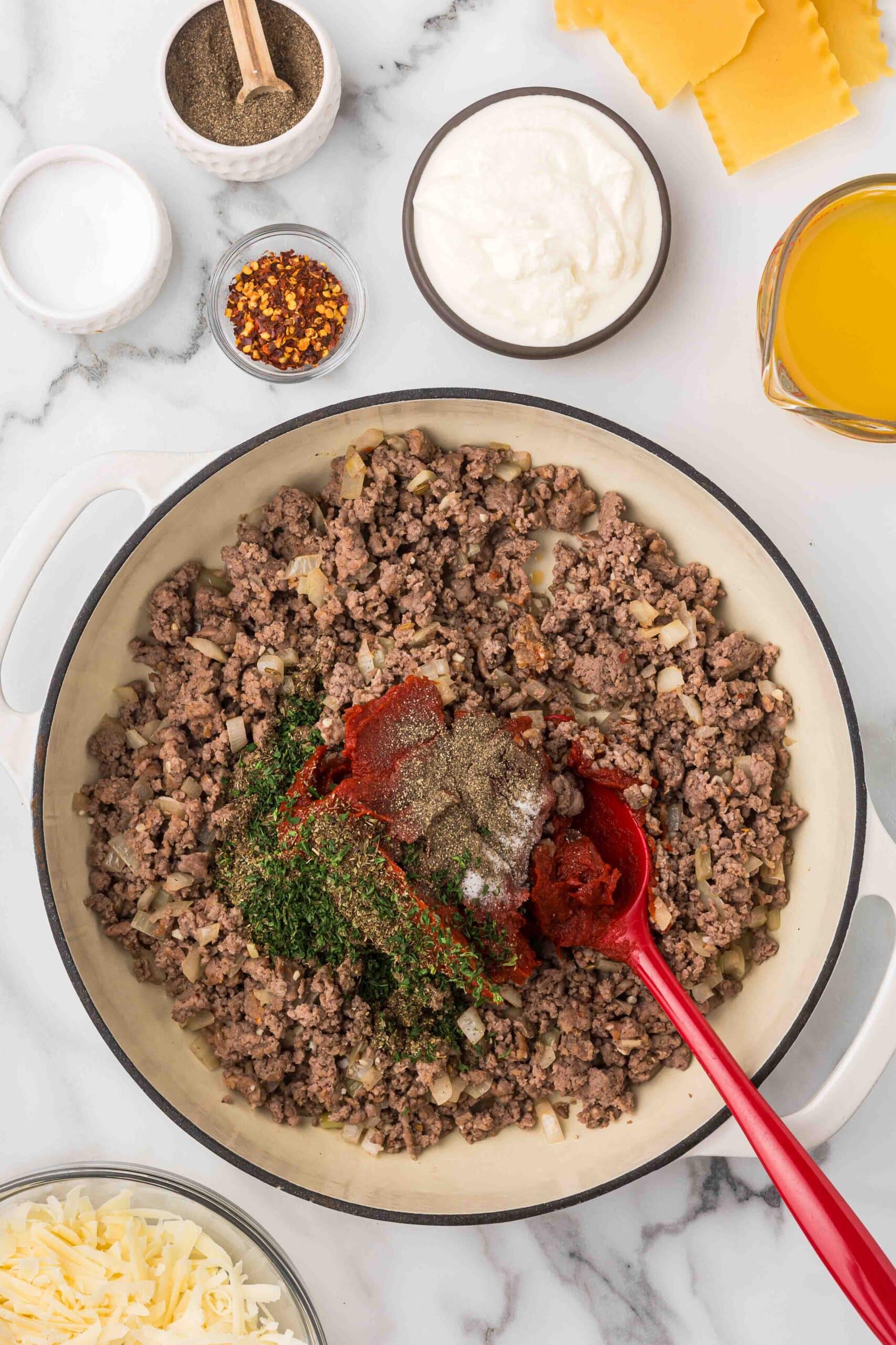 A white ceramic dish with ground meat, onions, tomato paste, and seasonings with a measuring cup of oil, a bowl of ricotta cheese, a bowl of red chili flakes, a bowl of salt, a bowl of pepper, and lasagna noodles, and a bowl of shredded mozzarella cheese. 