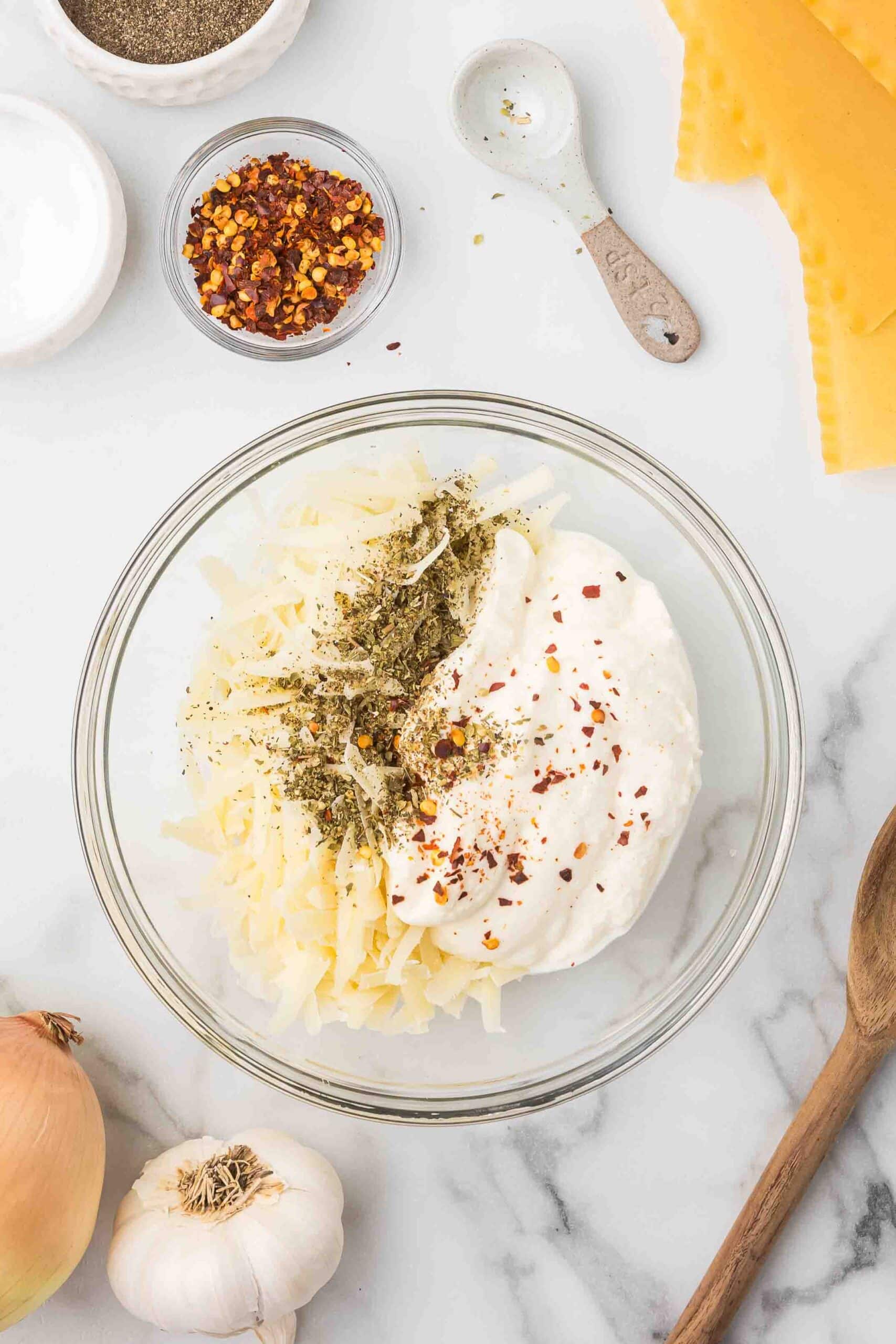 A bowl of pepper, a bowl of salt, a bowl of red chili flakes, an empty teaspoon, lasagna noodles, and a bowl of the cheese mixture, a wooden spoon, an onion and a bulb of garlic. 