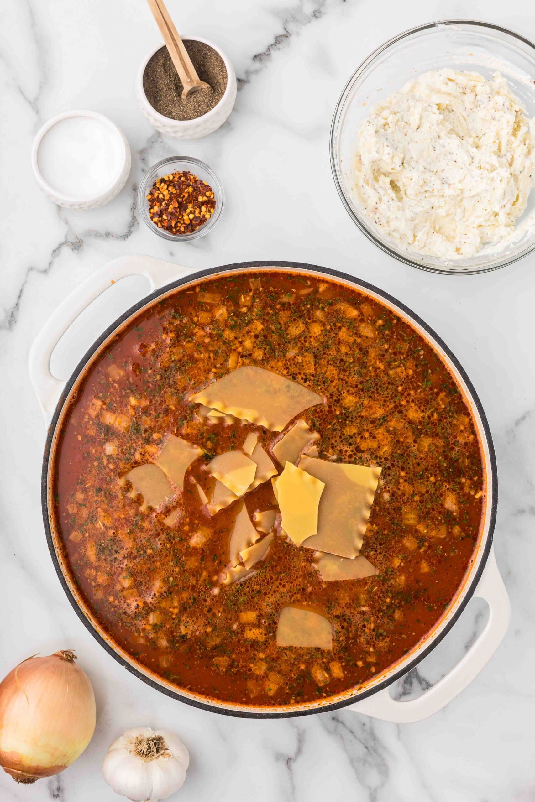 A ceramic dish with lasagna soup with lasagna noodles being added, a bowl of the cheese mixture, a bowl of red chili flakes, a bowl of salt, a bowl of pepper with a spoon in it, an onion and a bulb of garlic. 