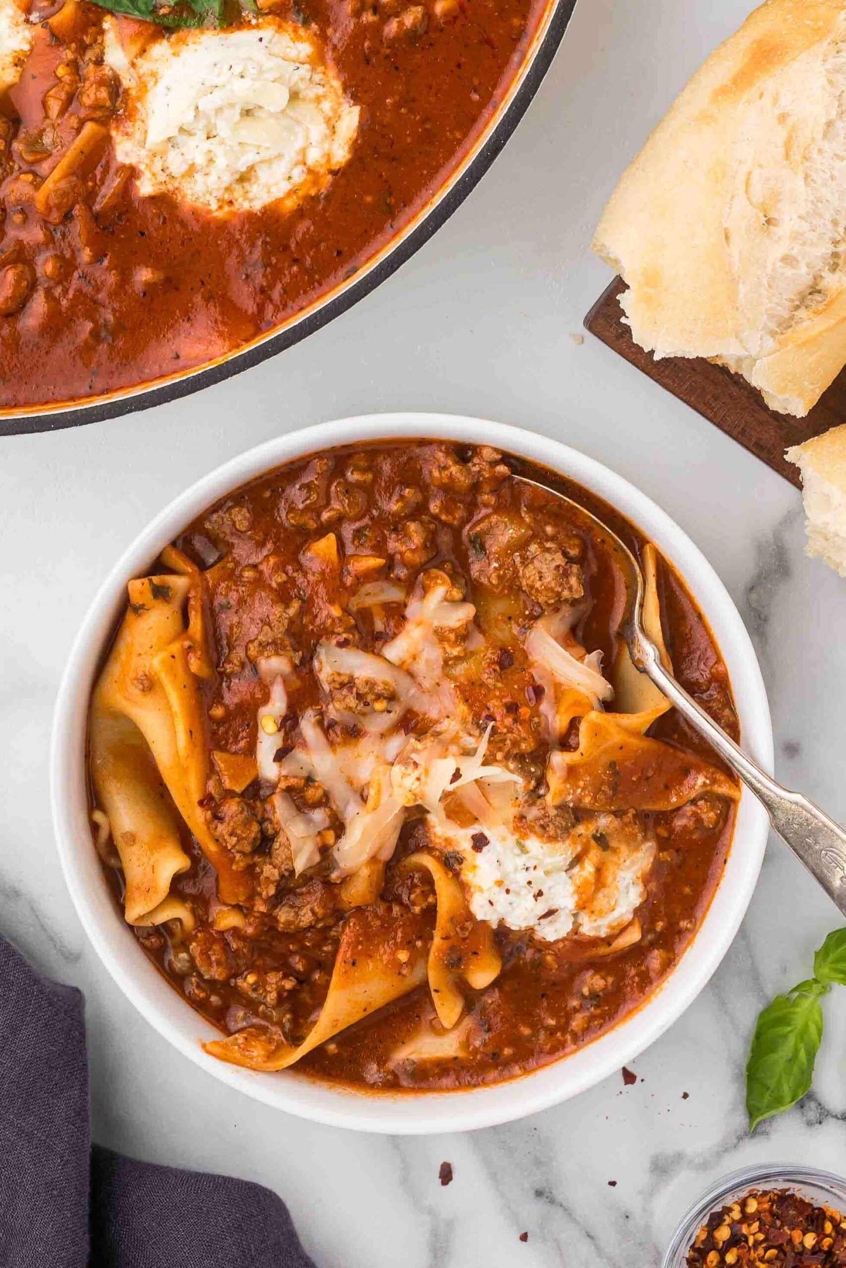 Lasagna soup in a large bowl and a small bowl with a spoon, a baguette on a wooden cutting board, basil leaves, a bowl of red chili flakes and a grey dish towel. 